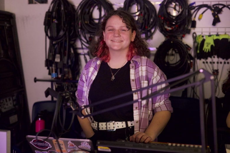 A student smiles in front of a stage production console. Behind them is a wall of wrapped cables and production gear.